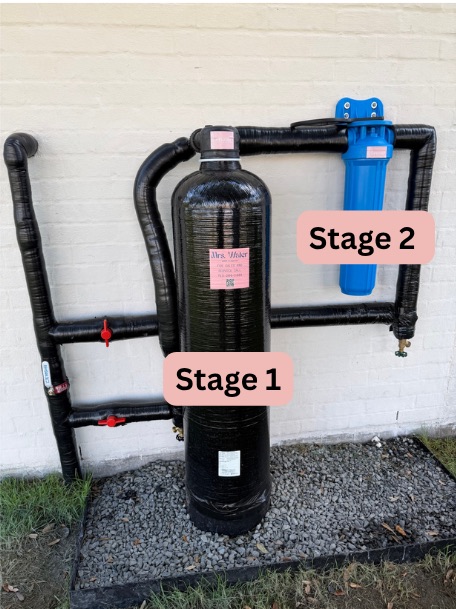 A Mrs. Water high-capacity filtration system installed against an exterior wall, showing the Stage 1 filtration tank and Stage 2 sediment filter with insulated piping.