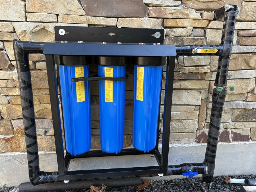 A three-stage Mrs. Water filtration system installed against a stone wall, with insulated piping and a bypass.